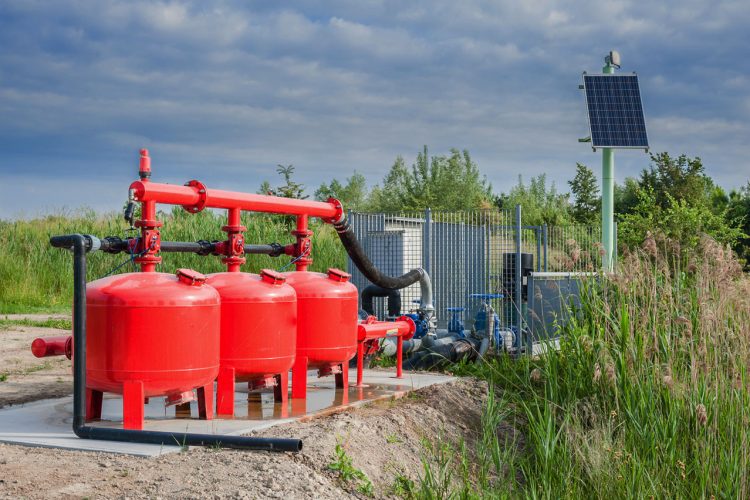 Image of a rural water well pump system featuring three large red tanks and connected piping. A solar panel is mounted on a pole nearby, indicating the system's use of renewable energy. The setup is situated outdoors with greenery and a cloudy sky in the background.